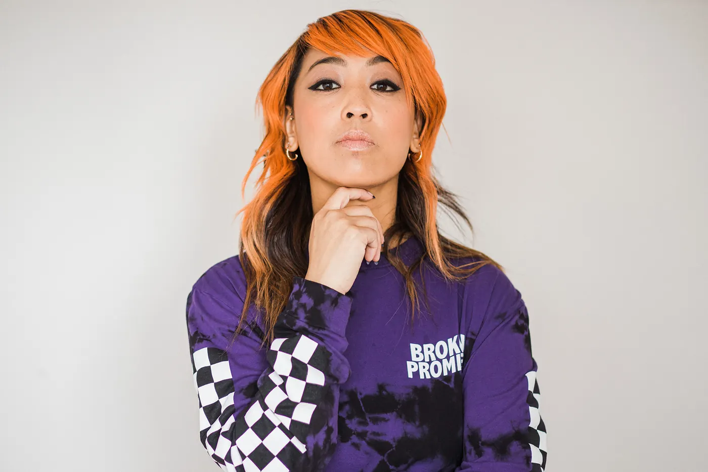 Cell7 with orange hair and a hand under her chin, in a purple tie-dye top with checkered sleeves