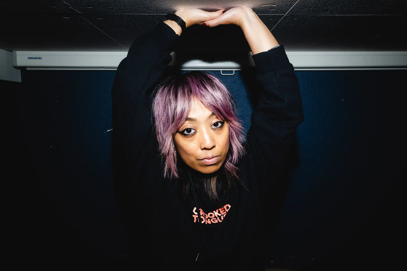 Cell7 with lilac hair and both arms raised above her head against a dark blue wall