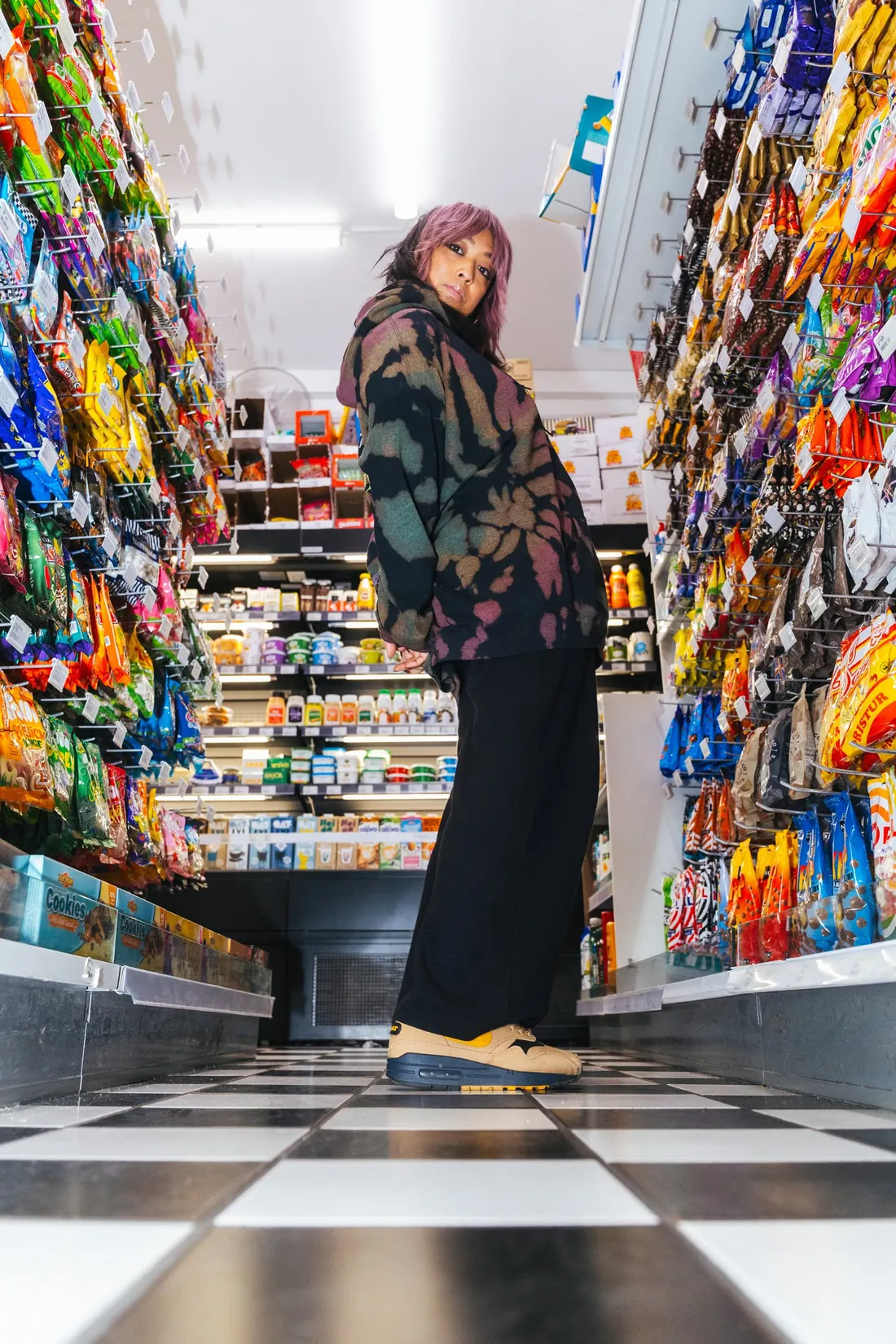 Cell7 in a tie-dye hoodie and wide black trousers, looking back over her shoulder in a candy-lined corner shop aisle with a checkered floor