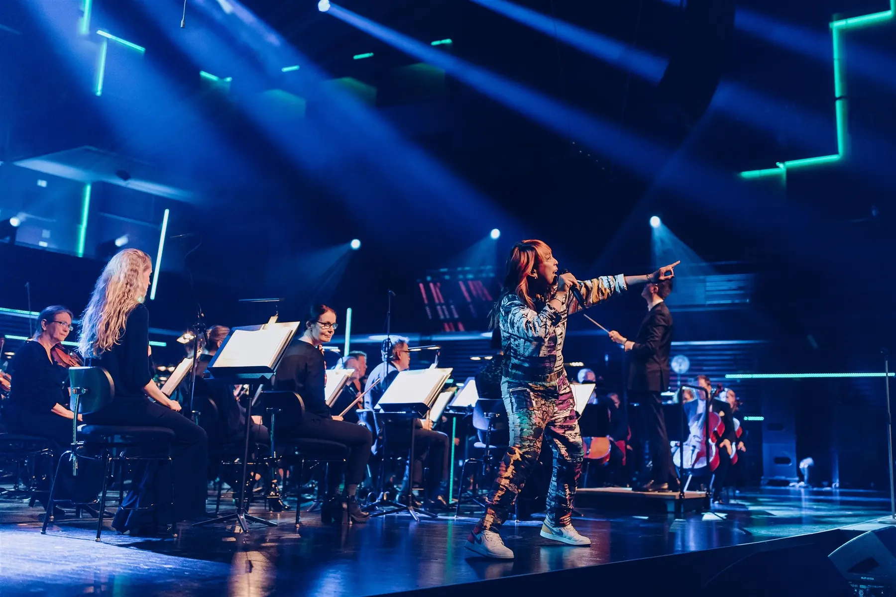 Cell7 rapping with a microphone on stage in front of an orchestra and conductor under blue stage light